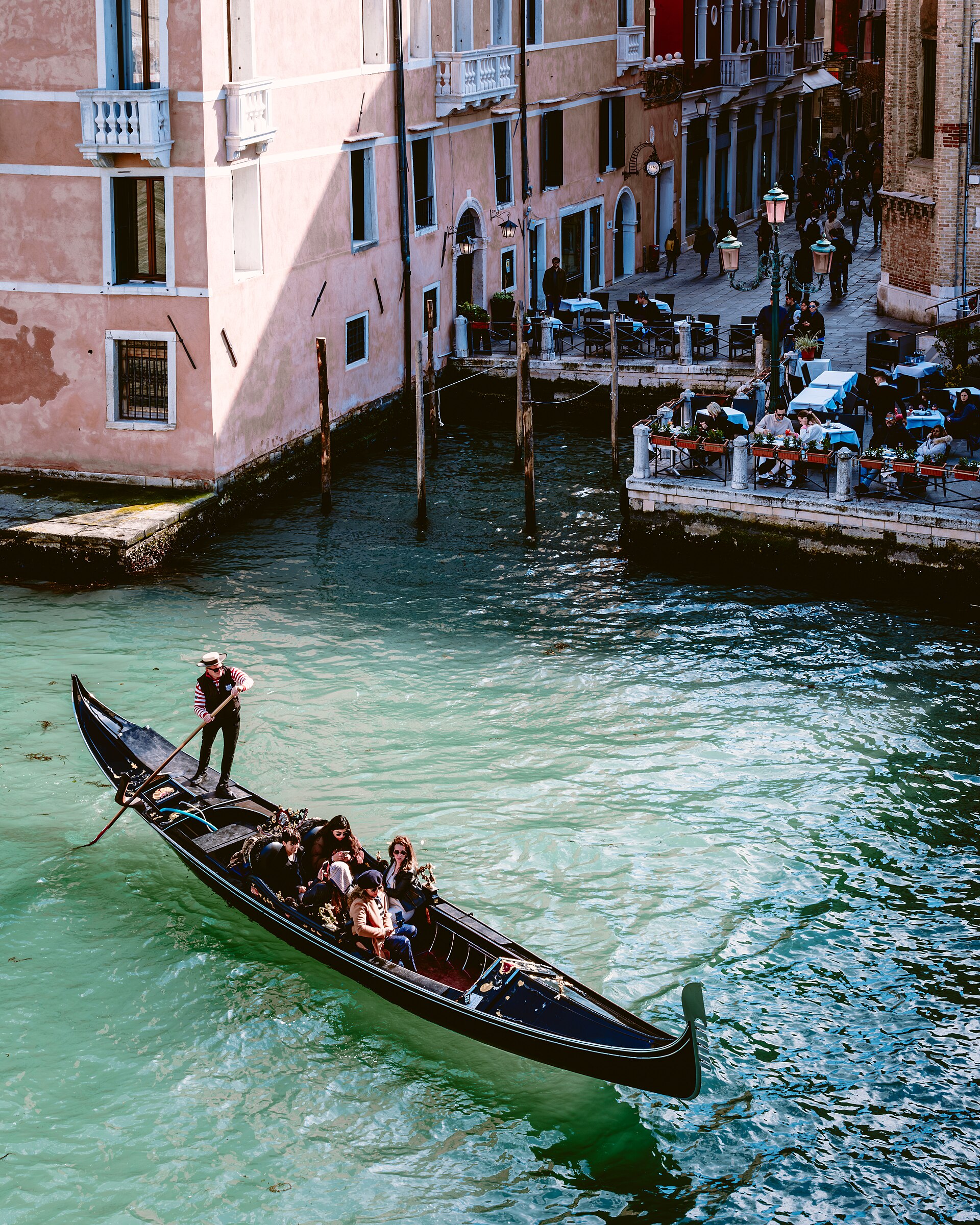 A gondolier uses one oar to guide a black gondola with passengers through a sunlit canal in Venice.